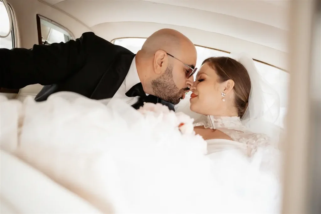 The bride and groom in a wedding dress are sitting close together in the back seat of a car and are about to kiss.