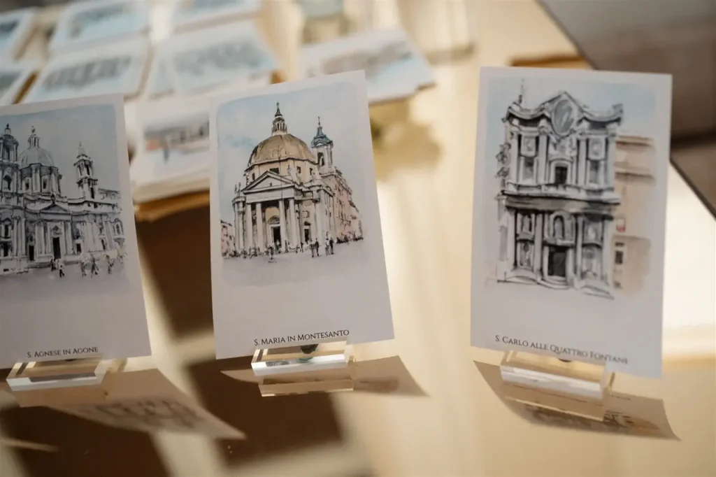 Three postcards displayed on holders show illustrations of classical buildings, including s. Maria in montesanto and s. Carlo alle quattro fontane, on a reflective surface.