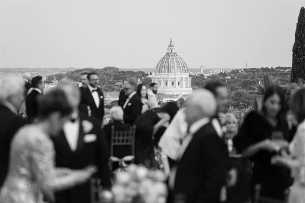 People in formal dress are at an outdoor event with the dome of st. Peter's basilica visible in the background. The scene is in black and white.
