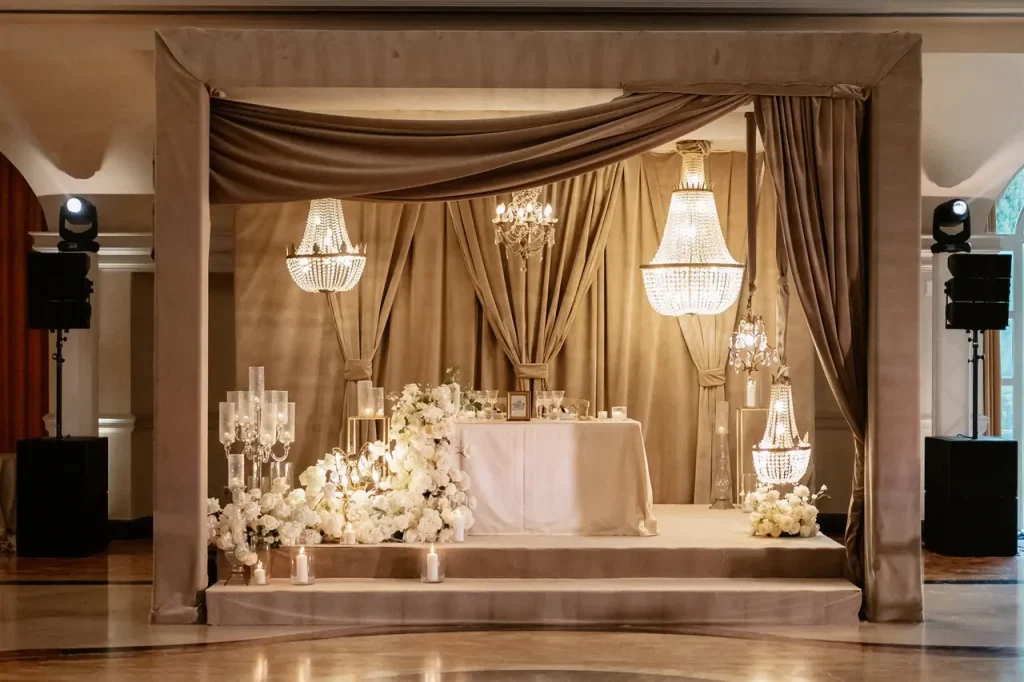 The elegant wedding stage at villa miani features draped fabrics, chandeliers, a white floral arrangement and lit candles surrounding a beautifully set table on the platform.