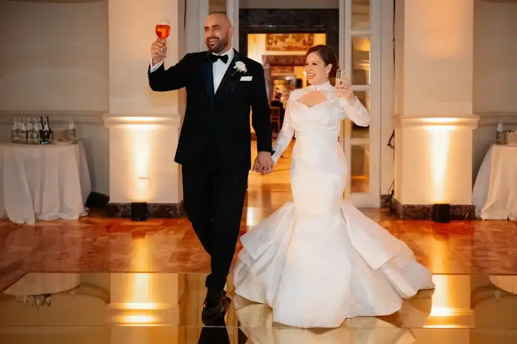 Villa miani: a bride and groom, holding drinks, smile as they enter a room. The groom wears a black suit and the bride a white dress. They hold hands and appear to celebrate.