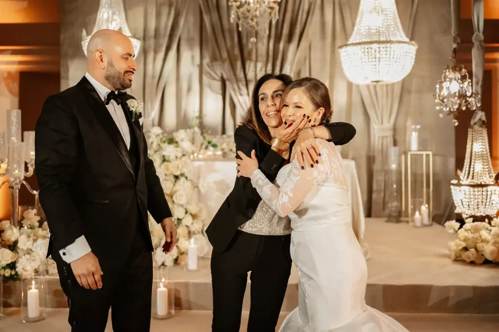 A bride in a white dress is embraced by a smiling woman in a suit, with a bald man in a dinner jacket standing nearby. In the background, flower arrangements and chandeliers.