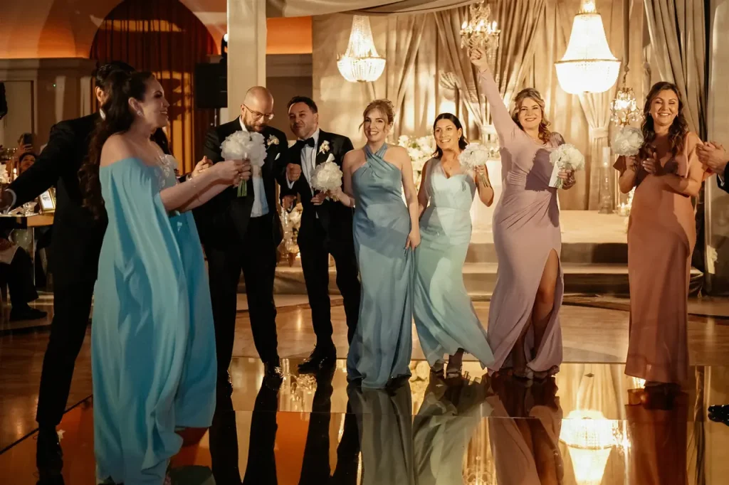 A group of people in formal dress stand and smile on a polished floor, with women holding bouquets. Chandeliers and elegant draperies are in the background.