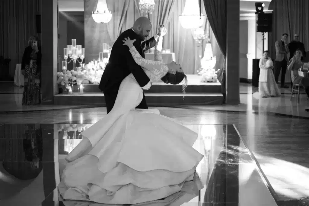 The bride and groom dance elegantly in a dimly lit venue, with chandeliers and draped curtains in the background.