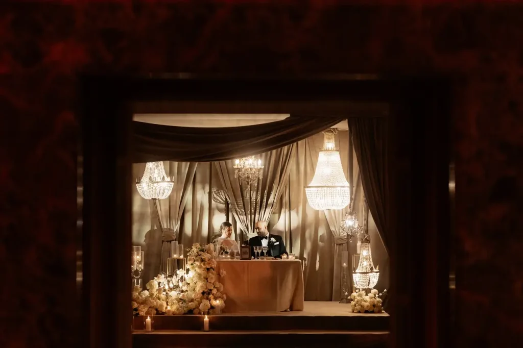 A couple is seated at an ornate table, under chandeliers on a stage, surrounded by draped fabrics and floral arrangements.