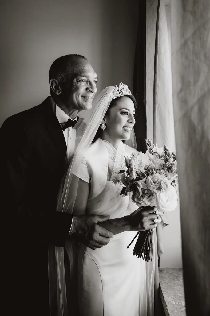 Vatican wedding: the story of gabriela and angel A bride in a wedding dress and veil holds a bouquet and is standing near a window of the hotel de la ville in rome, smiling, with an older man in a tuxedo behind her, both facing outward.