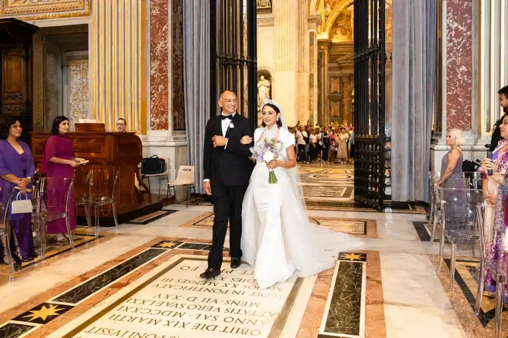 A bride in a white gown walks down the aisle with an elderly man in a suit inside the lavish “choir chapel” at the vatican, while guests look on from both sides.