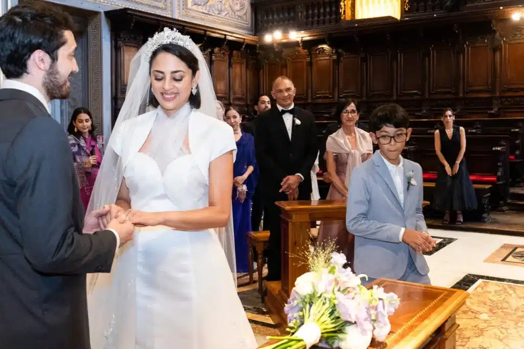 A married couple exchange rings at the altar in the choir chapel at the vatican, under the eyes of watching guests and with a bouquet on a table in the foreground.