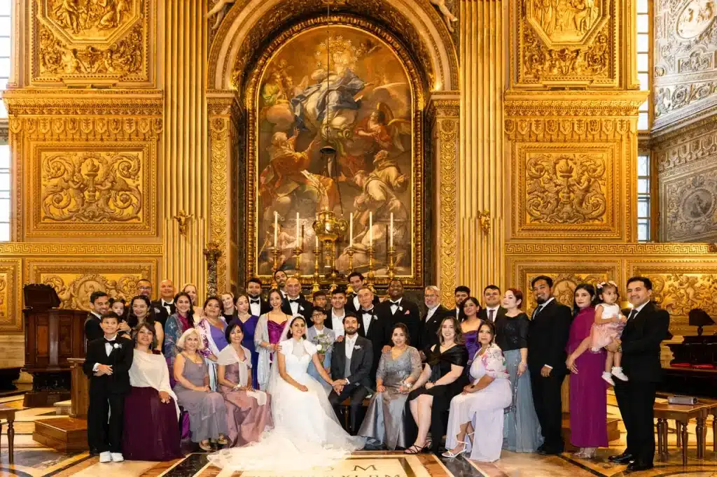 A large group of newlyweds pose in formal attire inside the lavish, gold-decorated “chapel of the choir” in the vatican, with a grandiose religious painting as a backdrop.