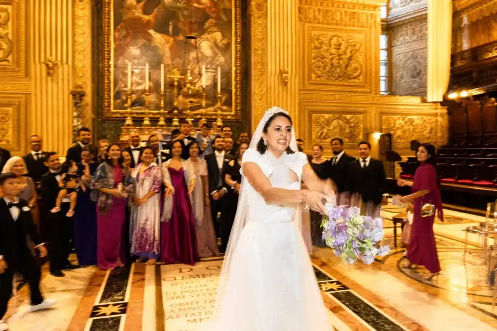 A bride in a white gown prepares to toss her bouquet as a group of wedding guests stand behind her in the large, ornate hall of the choir chapel at the vatican.