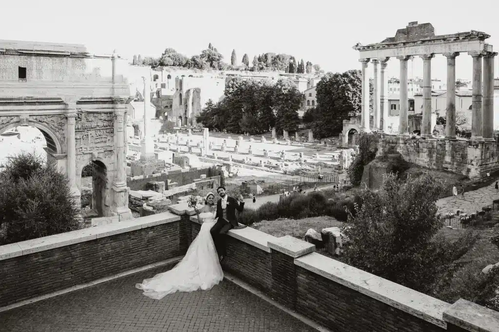 A married couple pose on a stone terrace overlooking ancient ruins, including columns and arches, after their romantic vatican wedding in this beautiful outdoor setting.