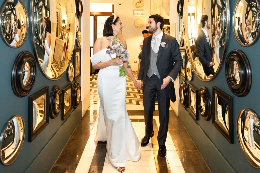 A married couple walk hand in hand down a hallway decorated with mirrors at rome's hotel de la ville, both dressed in formal wedding gowns and smiling after their vatican wedding.