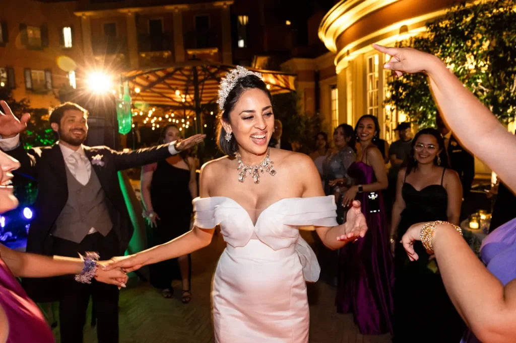 A bride in a white gown dances and smiles with guests during a lively, warmly lit indoor wedding reception at the elegant hotel de la ville in rome.