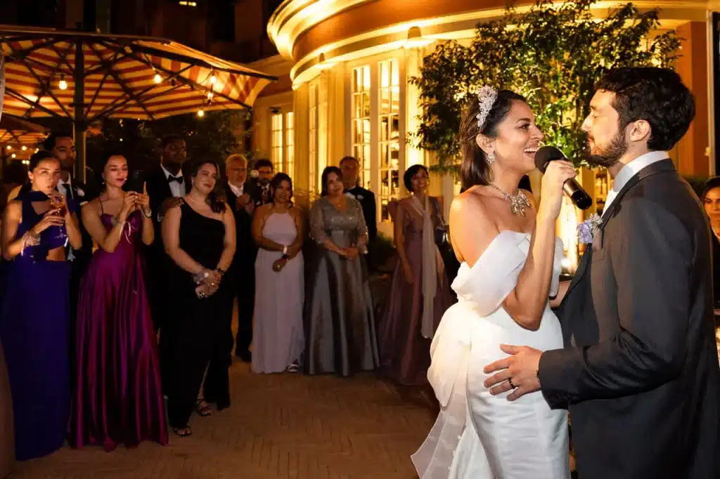 A bride in a white dress sings to her groom during an outdoor evening wedding reception near the hotel de la ville in rome, while guests watch and smile in the background.