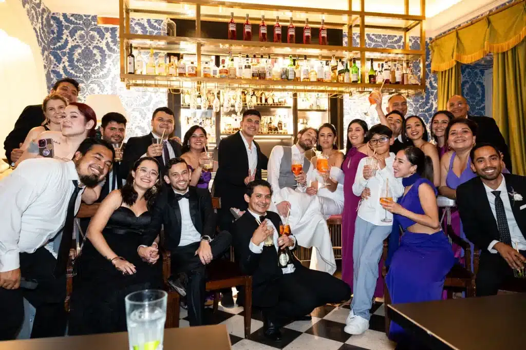 A large group of people dressed in formal attire pose together in an indoor setting, raising their glasses and smiling in front of a bar.